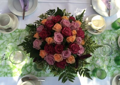 Blumengesteck aus pinken, roten und orangen Rosen, auf einem gedeckten Tisch.